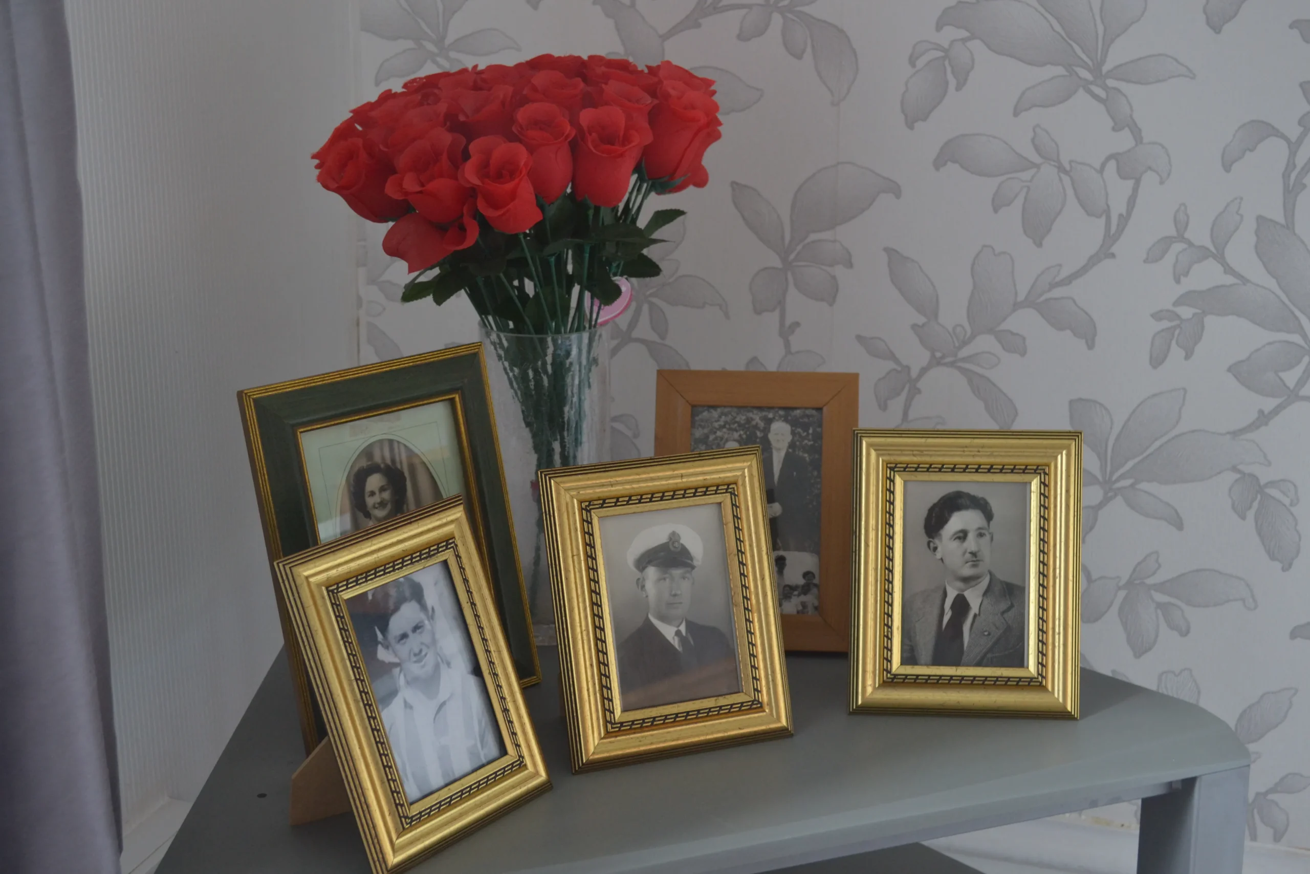 Framed family photographs and a vase of red roses on a table in a resident’s room at Chatsworth